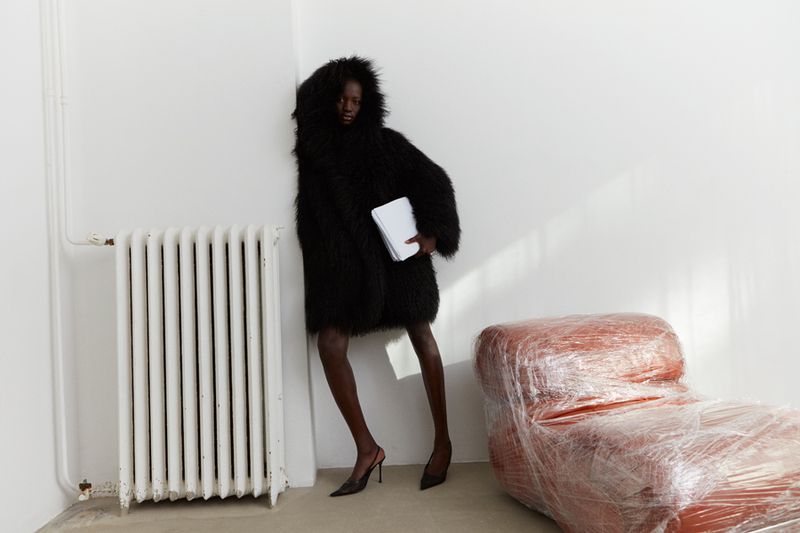 Fashion shot of a slender woman in a shaggy black coat holding papers, standing in a white corner with a radiator and wrapped couch.