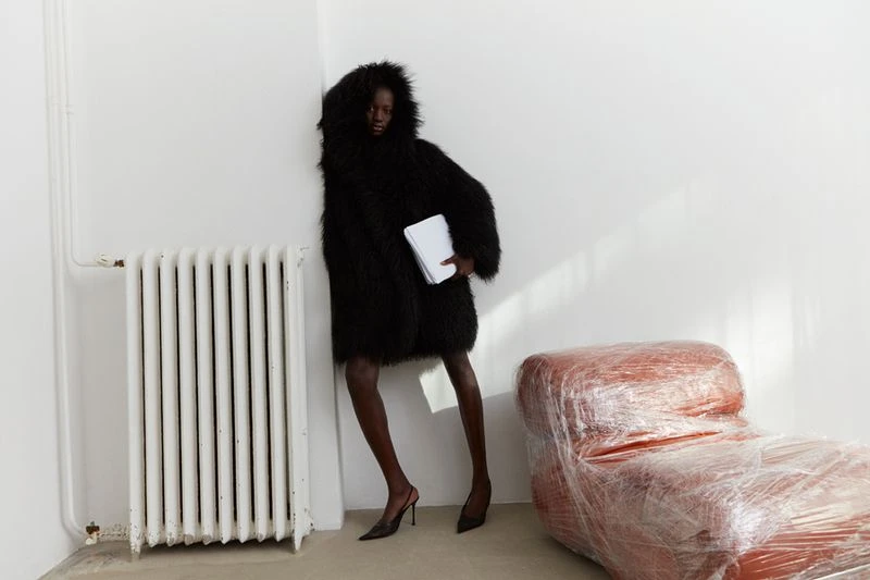 Fashion shot of a slender woman in a shaggy black coat holding papers, standing in a white corner with a radiator and wrapped couch.