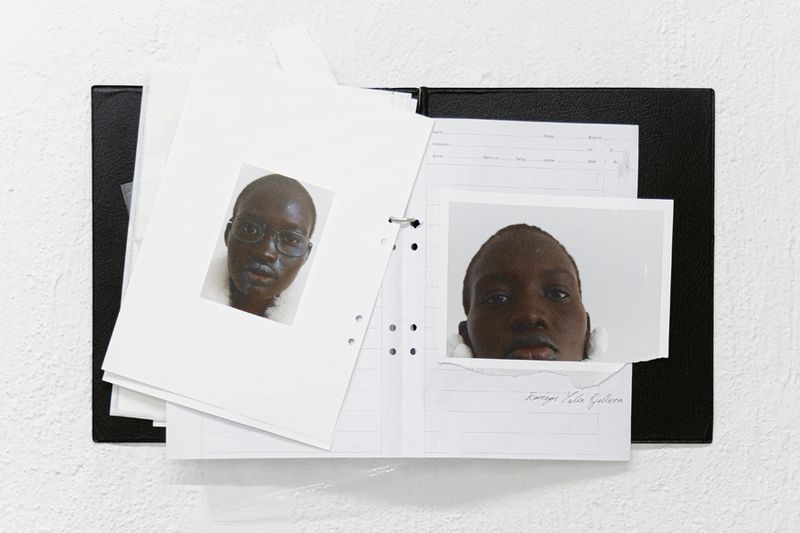 Photographs of an adult with dark skin, one with glasses, affixed to loose-leaf pages held by a black three-ring binder on white.