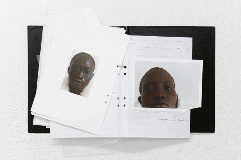 Photographs of an adult with dark skin, one with glasses, affixed to loose-leaf pages held by a black three-ring binder on white.