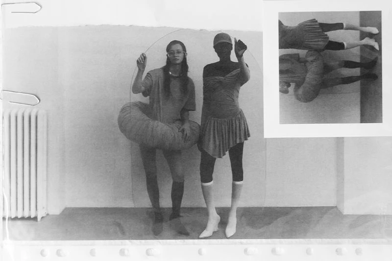 Black and white photograph featuring two adult women in a bright, indoor space, accompanied by a framed portrait and visible radiator.