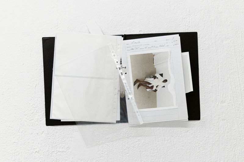 Black binder with pages and a photograph showing a seated person in a bright room against a white textured wall background.