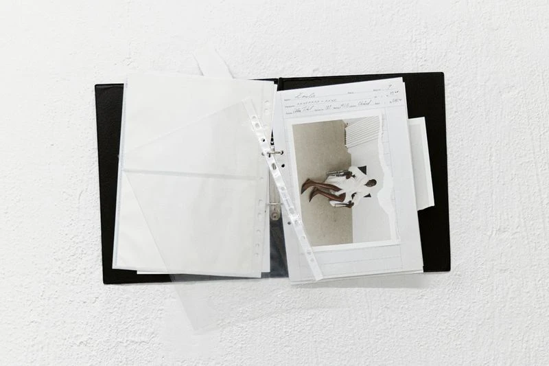 Black binder with pages and a photograph showing a seated person in a bright room against a white textured wall background.
