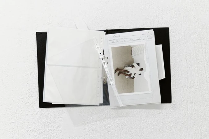 Black binder with pages and a photograph showing a seated person in a bright room against a white textured wall background.