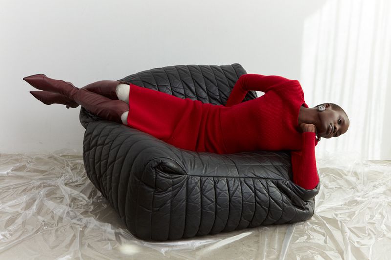 An adult female model, wearing a red dress and burgundy boots, is reclining on a black leather sofa with a relaxed expression.