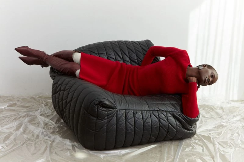 An adult female model, wearing a red dress and burgundy boots, is reclining on a black leather sofa with a relaxed expression.