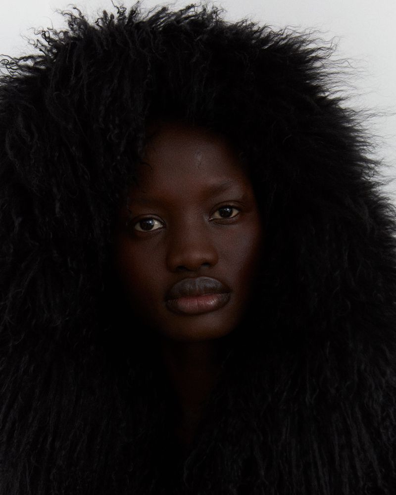 Close-up portrait of an adult Black woman with dark eyes and full lips, framed by dark, voluminous, curly hair against a plain background.
