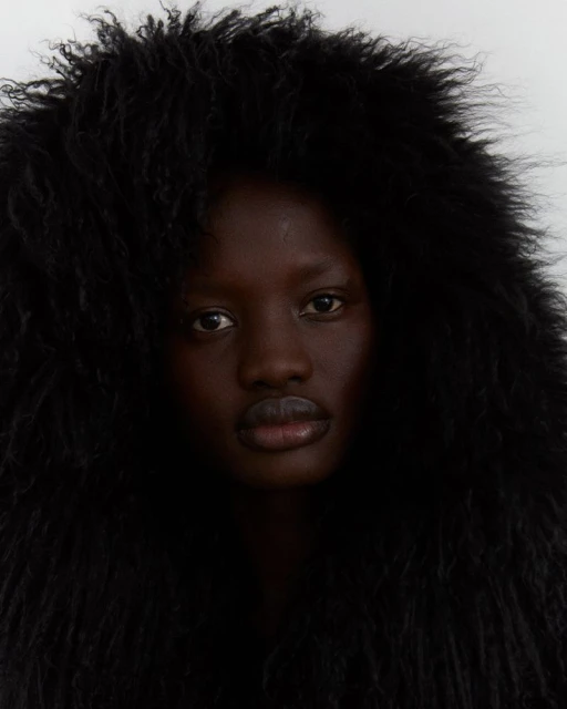 Close-up portrait of an adult Black woman with dark eyes and full lips, framed by dark, voluminous, curly hair against a plain background.