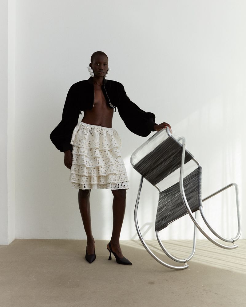 Fashion shot featuring a black female model wearing a layered white skirt, black jacket, earrings, heels and posing near a chair.