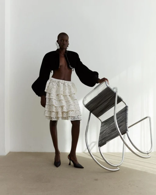 Fashion shot featuring a black female model wearing a layered white skirt, black jacket, earrings, heels and posing near a chair.