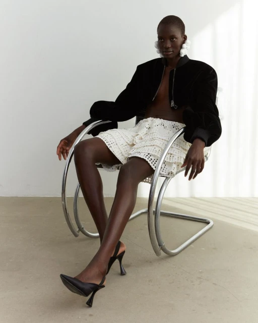 A dark-skinned woman in a black jacket and white shorts is seated in a metal chair against a white backdrop. She's wearing black heels.