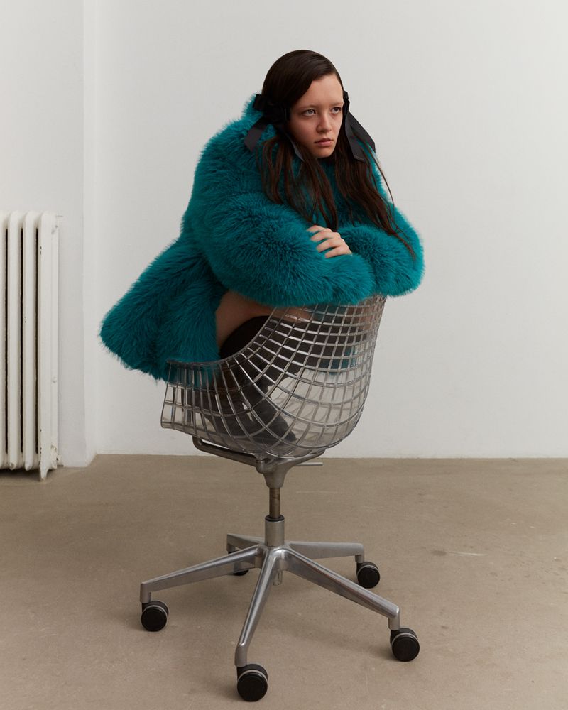 A solitary young adult female sits in a chrome office chair, wearing a textured teal fur coat, and looking off-camera in a white room.