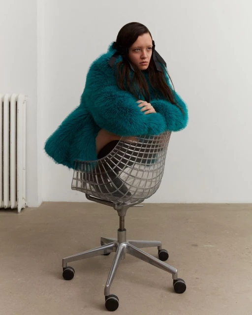 A solitary young adult female sits in a chrome office chair, wearing a textured teal fur coat, and looking off-camera in a white room.