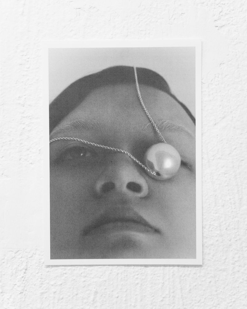 Monochromatic portrait of a person's face partially obscured by a large pearl on a delicate chain against a textured backdrop.
