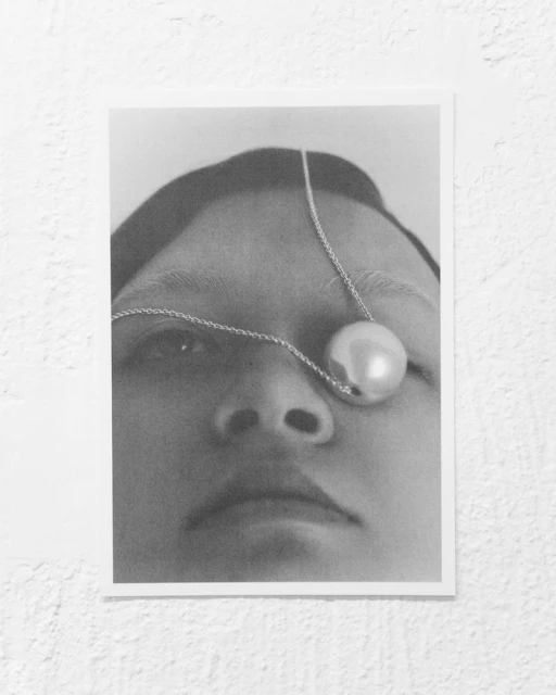 Monochromatic portrait of a person's face partially obscured by a large pearl on a delicate chain against a textured backdrop.