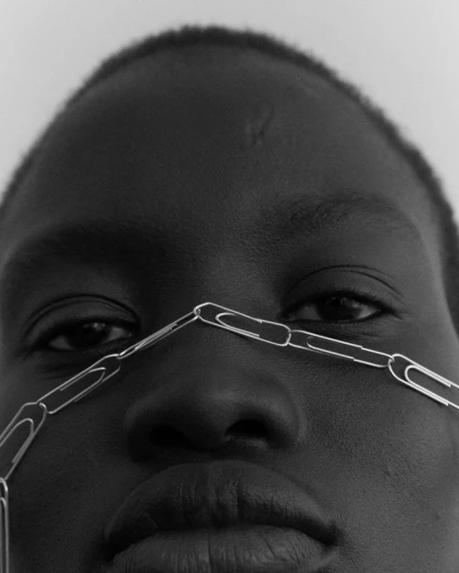 Striking close-up studio portrait of a Black adult female with paper clips around her nose. The image is black and white.
