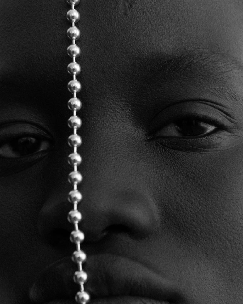 Close-up portrait of a Black adult female with dark eyes, a silver ball chain, and diffused lighting in a studio setting.