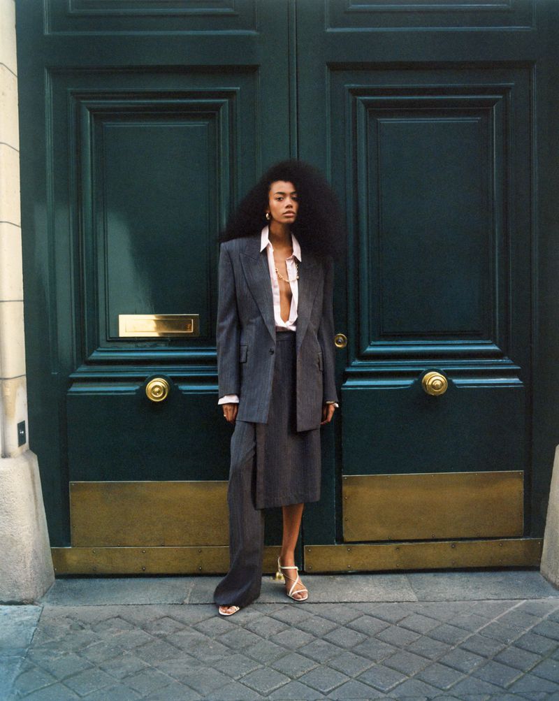 Adult woman with Afro hairstyle poses in gray skirt suit in front of a large green door with gold accents and cobblestone ground.