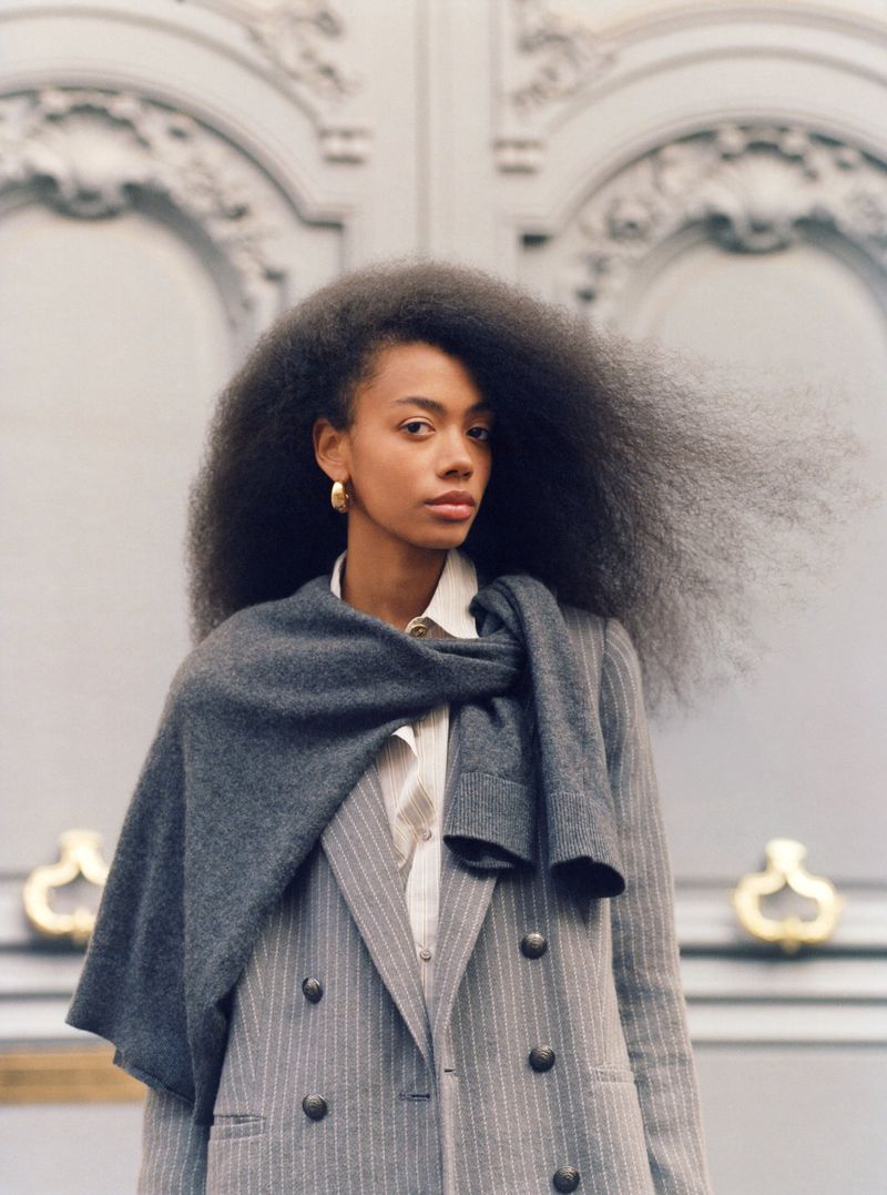 Portrait of a stylish African American woman with voluminous afro hair, wearing a grey suit and scarf in front of grey ornate doors.