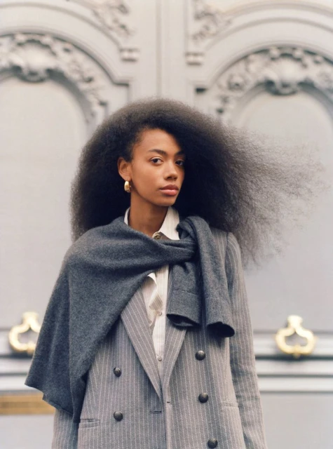 Portrait of a stylish African American woman with voluminous afro hair, wearing a grey suit and scarf in front of grey ornate doors.