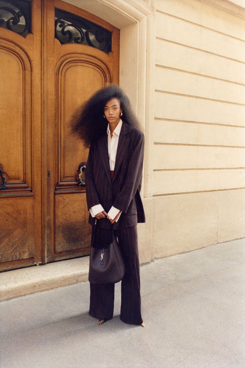 Elegant Black woman with large Afro in a pinstripe suit and large leather purse standing in front of wooden doors on a city street.