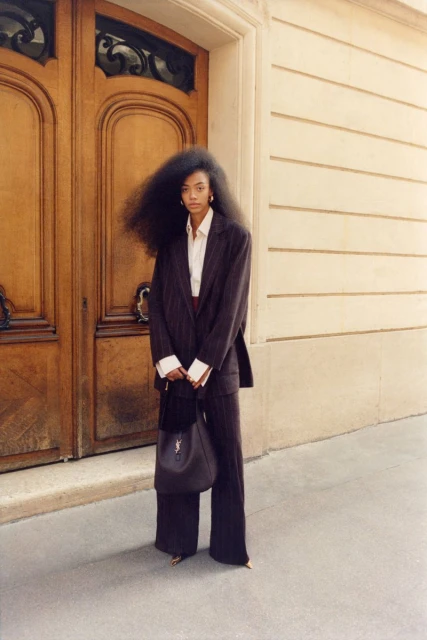 Elegant Black woman with large Afro in a pinstripe suit and large leather purse standing in front of wooden doors on a city street.