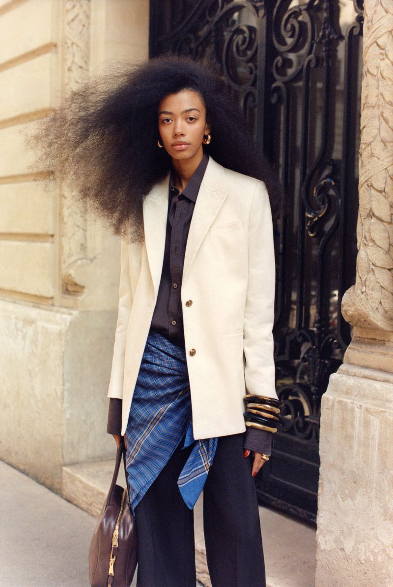 Portrait of a Black woman with an afro wearing a white blazer and dark pants, posing outdoors in front of a decorative wrought iron gate.