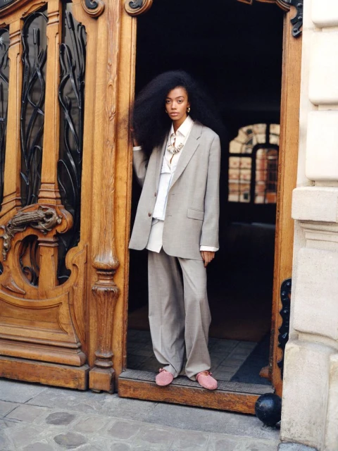 An adult female with large afro hair, wearing a grey suit and pink shoes, stands in a doorway, posing in front of an ornate wooden door.
