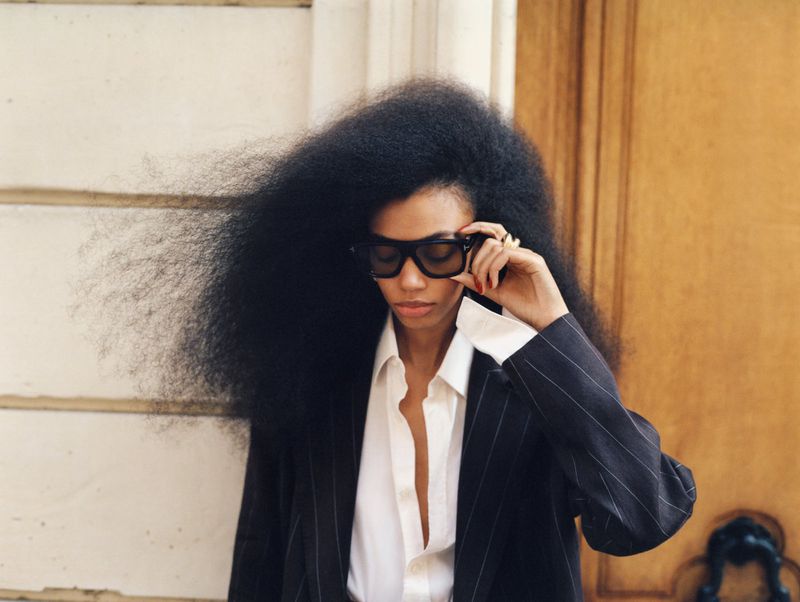 Portrait of an adult woman with oversized afro hairstyle in pinstripe suit and sunglasses against a white wall, fashion-forward and stylish.