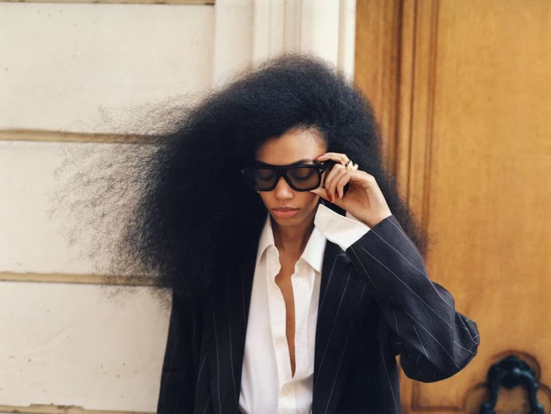 Portrait of an adult woman with oversized afro hairstyle in pinstripe suit and sunglasses against a white wall, fashion-forward and stylish.