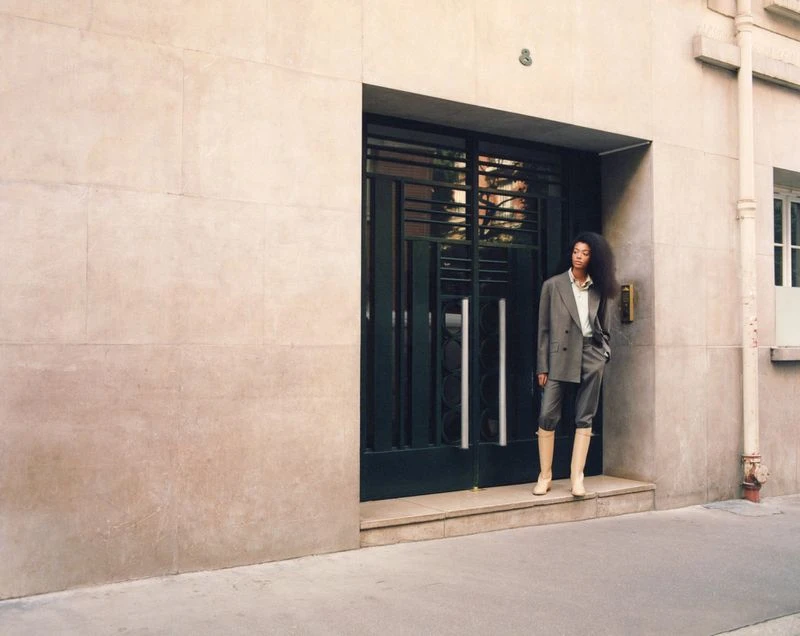 An adult female poses outside a building in a grey suit and tall boots. The setting is an urban environment with a neutral palette.