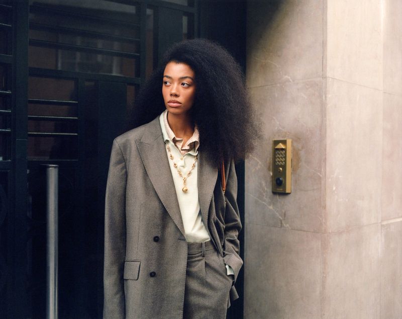 Woman in a grey pant suit with a gold necklace and afro stands in front of a textured wall with a black iron door.