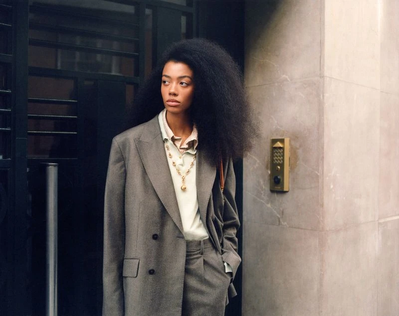 Woman in a grey pant suit with a gold necklace and afro stands in front of a textured wall with a black iron door.