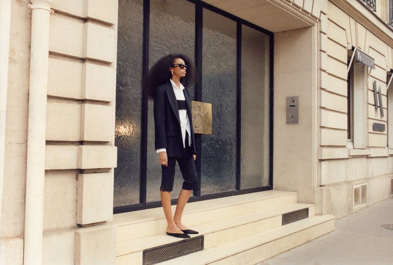 Elegant adult woman in a stylish black suit posing in front of a modern stone building with large glass doors and Parisian architecture.