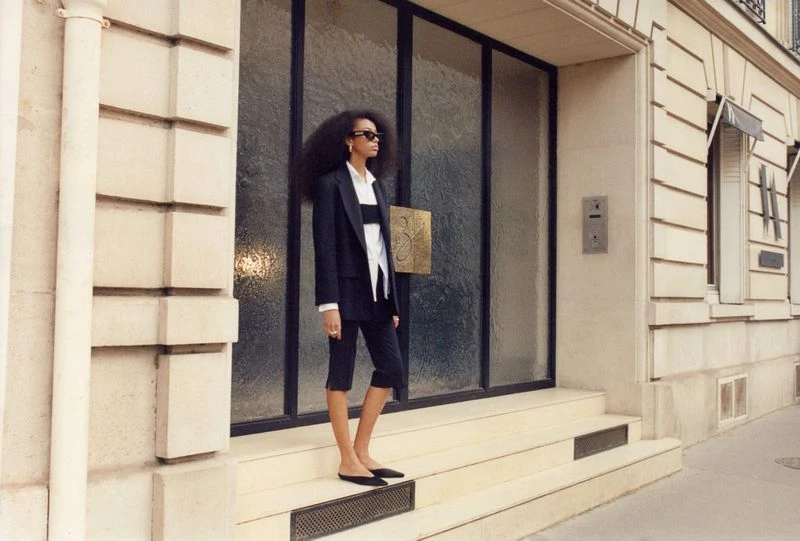 Elegant adult woman in a stylish black suit posing in front of a modern stone building with large glass doors and Parisian architecture.