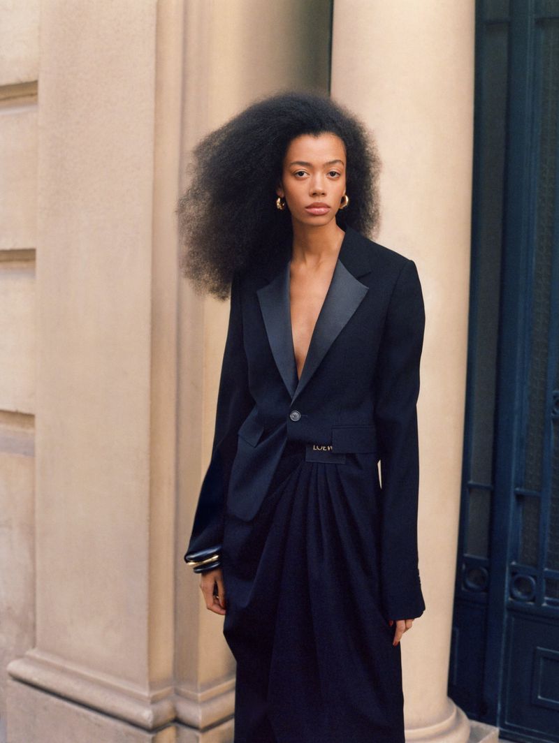 A stylish adult woman with afro-style hair wears a black tuxedo blazer and dark skirt, posing outdoors against a neutral architectural background.