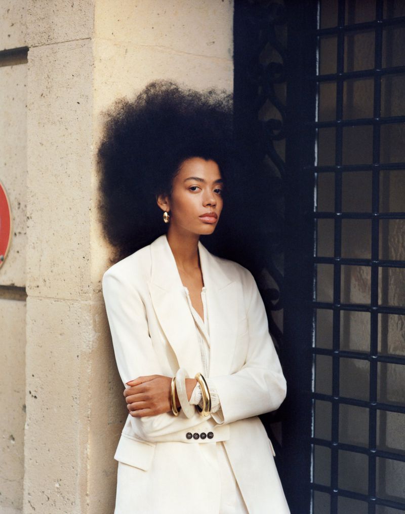 Portrait of a stylish adult woman in an off-white suit standing against a wall and doorway with a curly afro hairstyle.