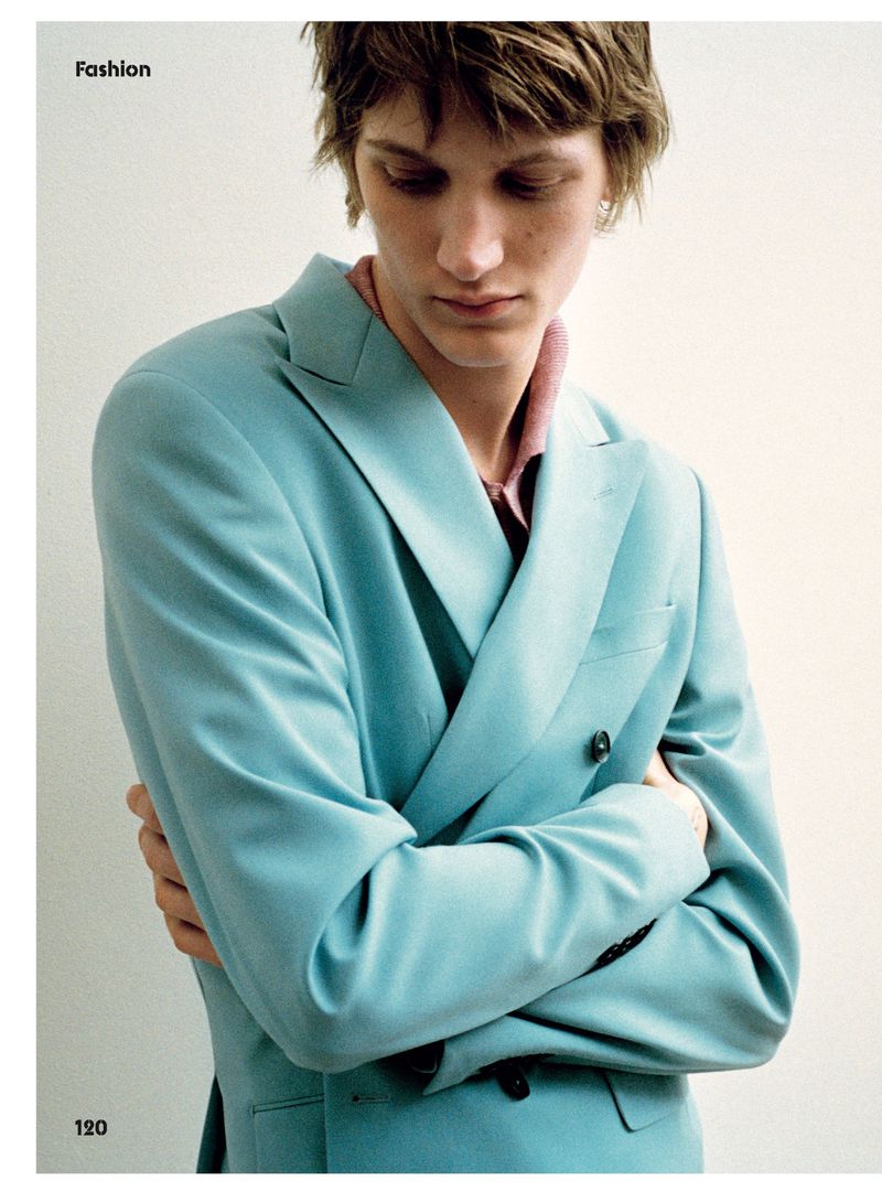Close-up shot of a blonde male fashion model in a light blue double-breasted suit looking down with his arms crossed against a white background.