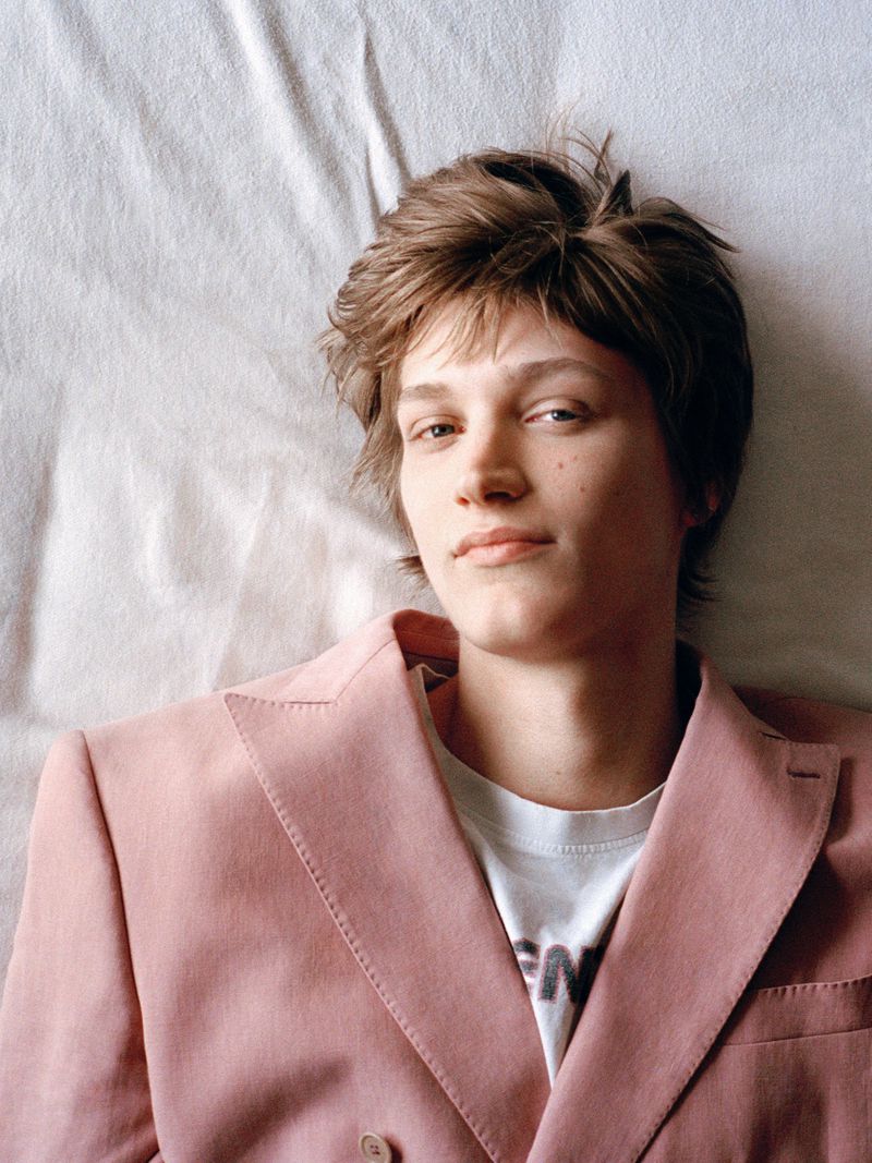 Portrait of a young adult man with light brown hair lying on a white surface wearing a pink double-breasted blazer and a white t-shirt.