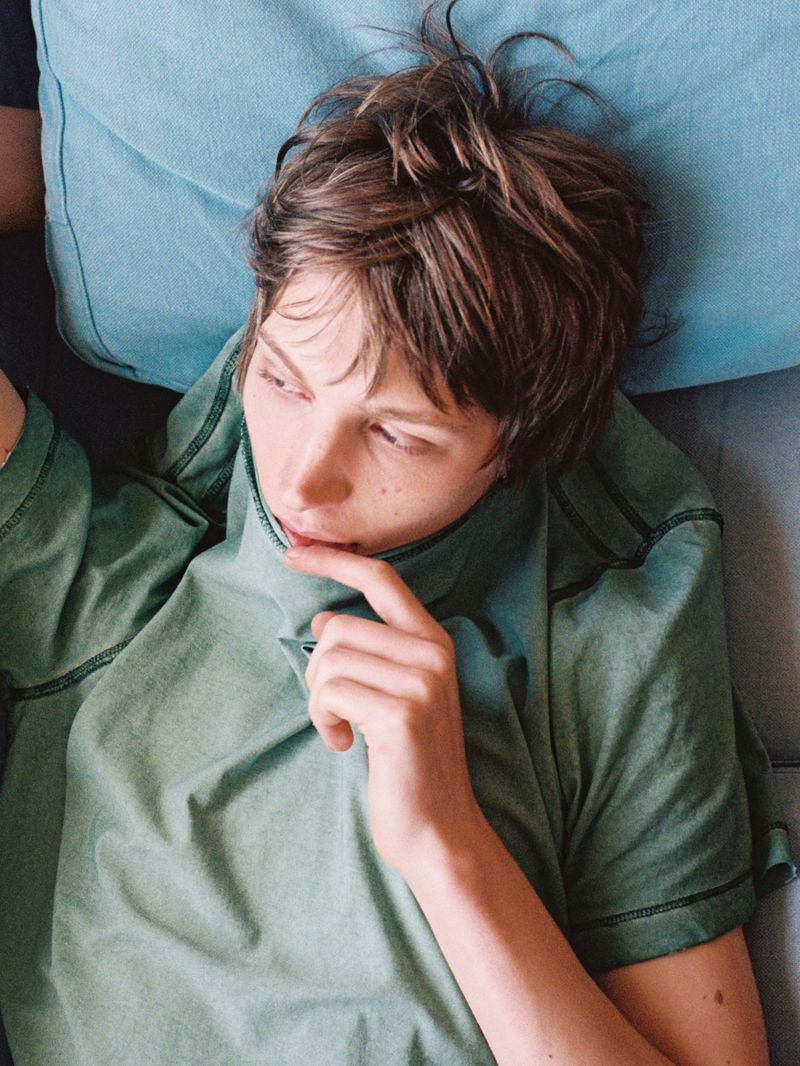 Person with short brown hair lying on a light blue pillow and wearing a green shirt, looking off to the side, hand near face.