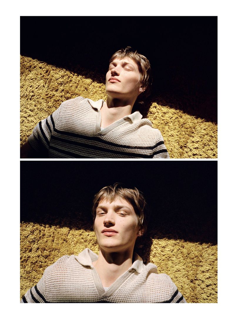 Male model lying on a textured yellow surface, wearing a collared shirt with black stripes and basking in natural light. Editorial fashion.