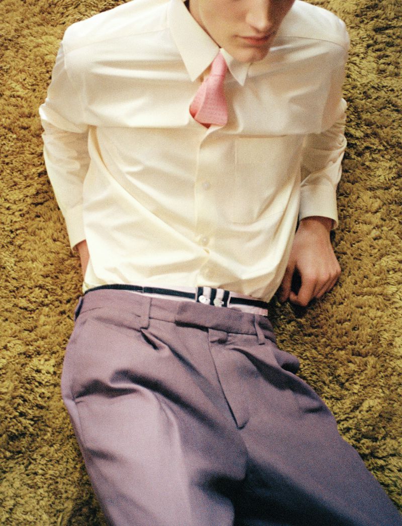 Young man lies on shag carpet wearing lavender pants, white dress shirt, and pink tie in a fashion or editorial style photograph.