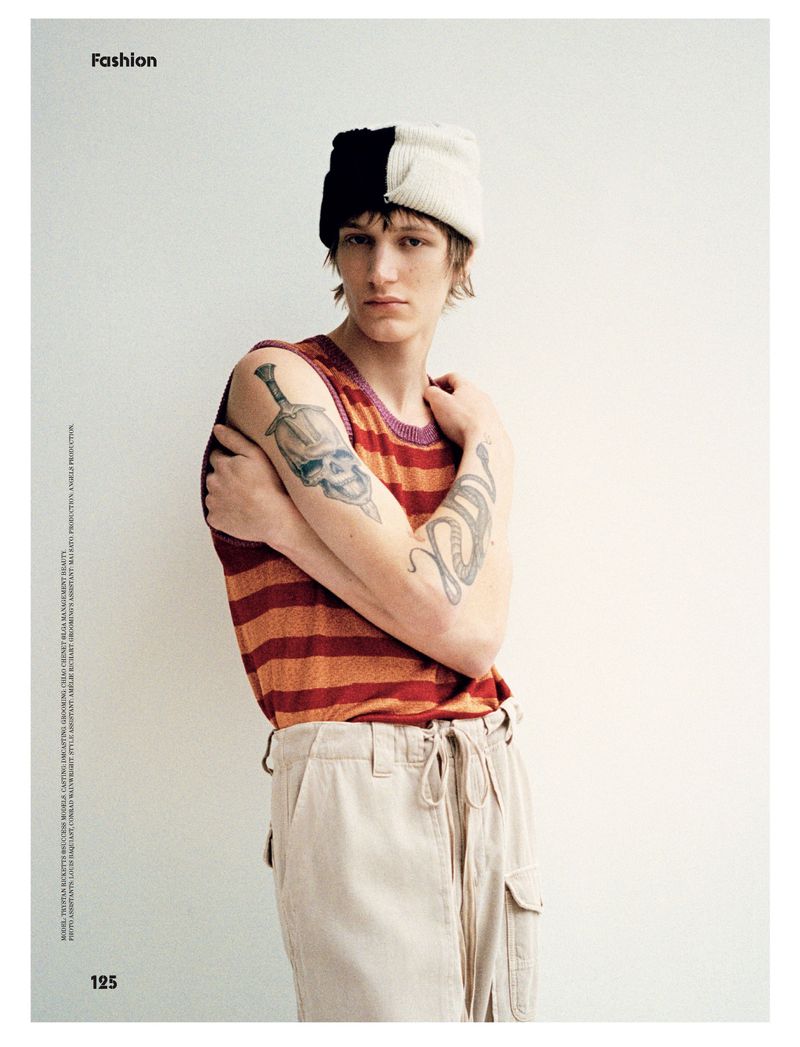 Fashion shot of a slender young man with tattoos, wearing a striped top and light pants, posing in front of a plain off-white wall.