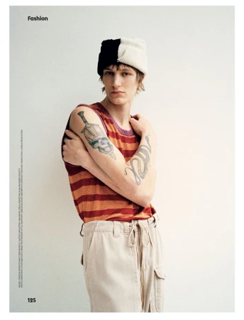 Fashion shot of a slender young man with tattoos, wearing a striped top and light pants, posing in front of a plain off-white wall.