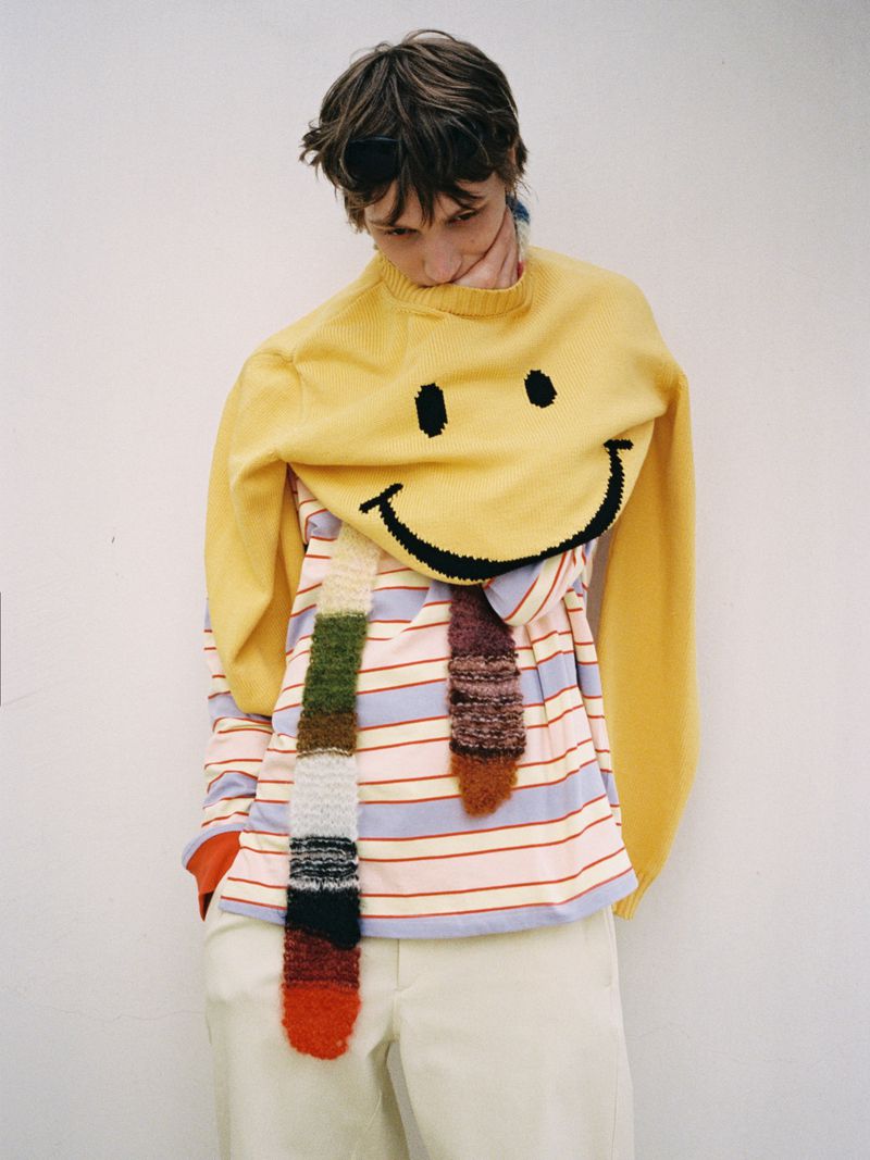 Model wearing a yellow smiley face sweater and striped long sleeve top against a white seamless background