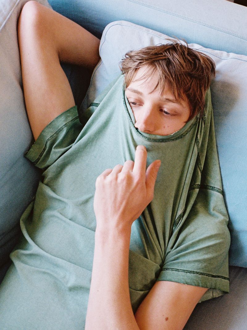 Young male model lying on a light blue sofa with a sage green t-shirt pulled up over his lower face