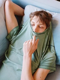 Young male model lying on a light blue sofa with a sage green t-shirt pulled up over his lower face