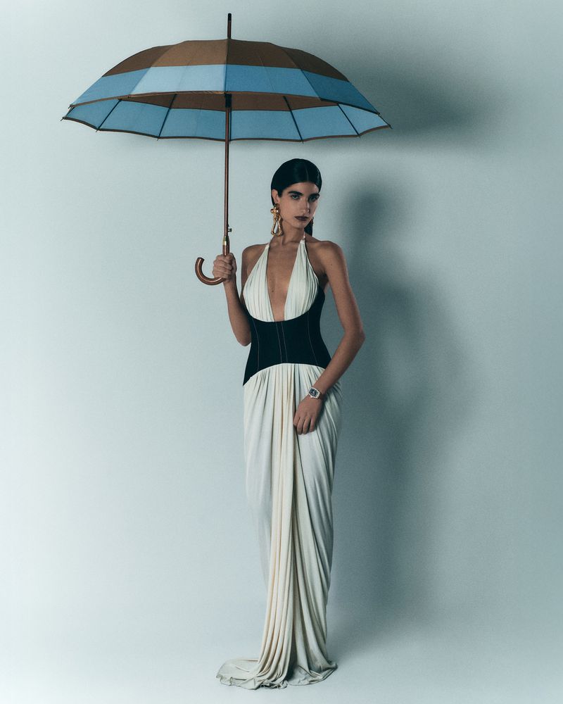 Elegant woman in a long white halter dress stands under a striped umbrella, accessorized with large gold earrings against a light backdrop.