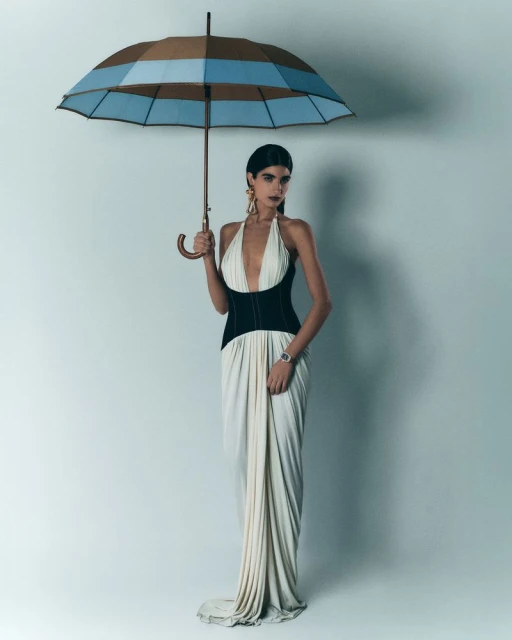 Elegant woman in a long white halter dress stands under a striped umbrella, accessorized with large gold earrings against a light backdrop.