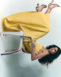 Woman wearing a bright yellow dress reclining backwards on a light grey plastic chair against a plain backdrop.